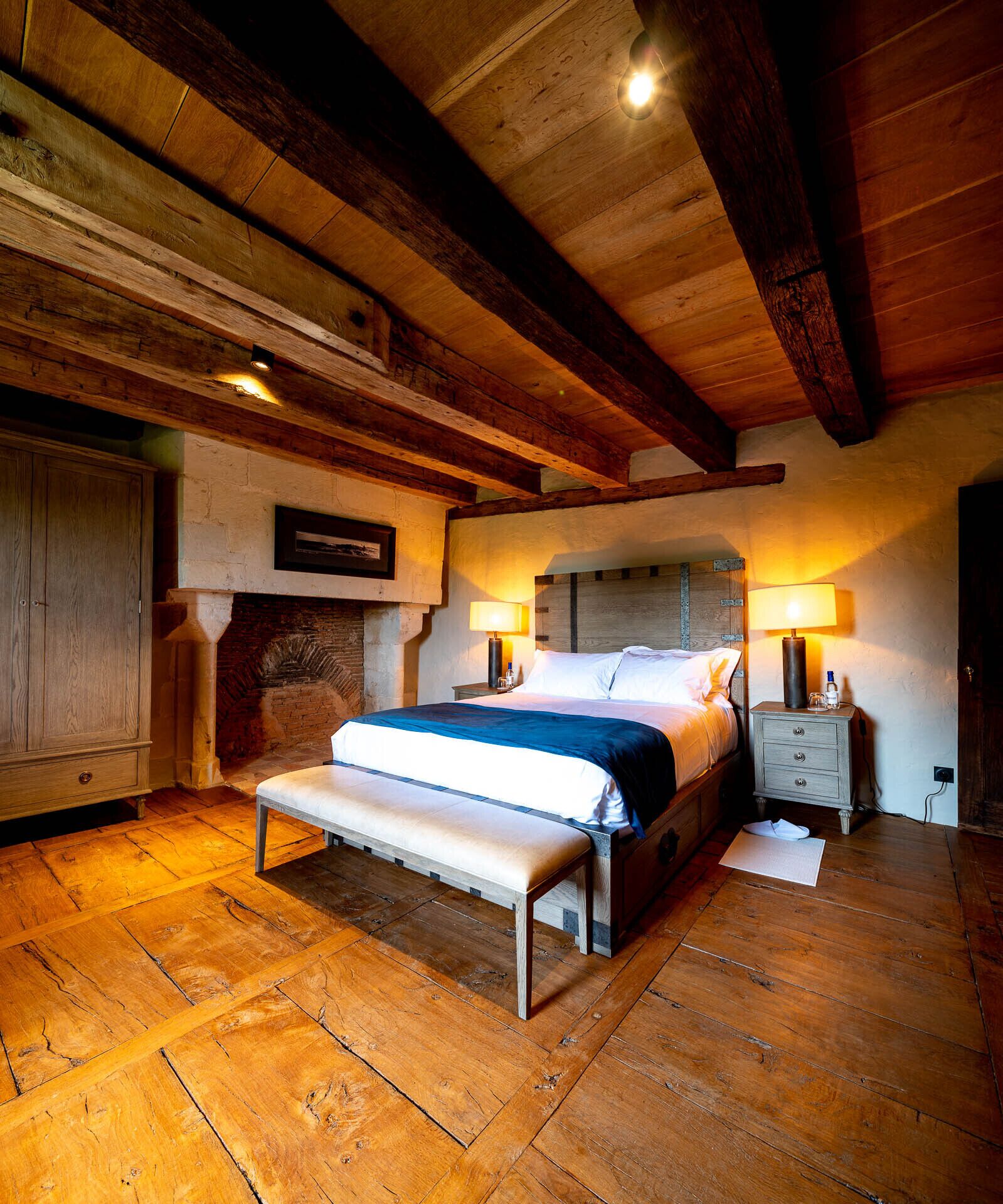 Une chambre confortable avec un plancher en bois, un lit recouvert d’une couverture bleue, un banc et une table de chevet avec une lampe.