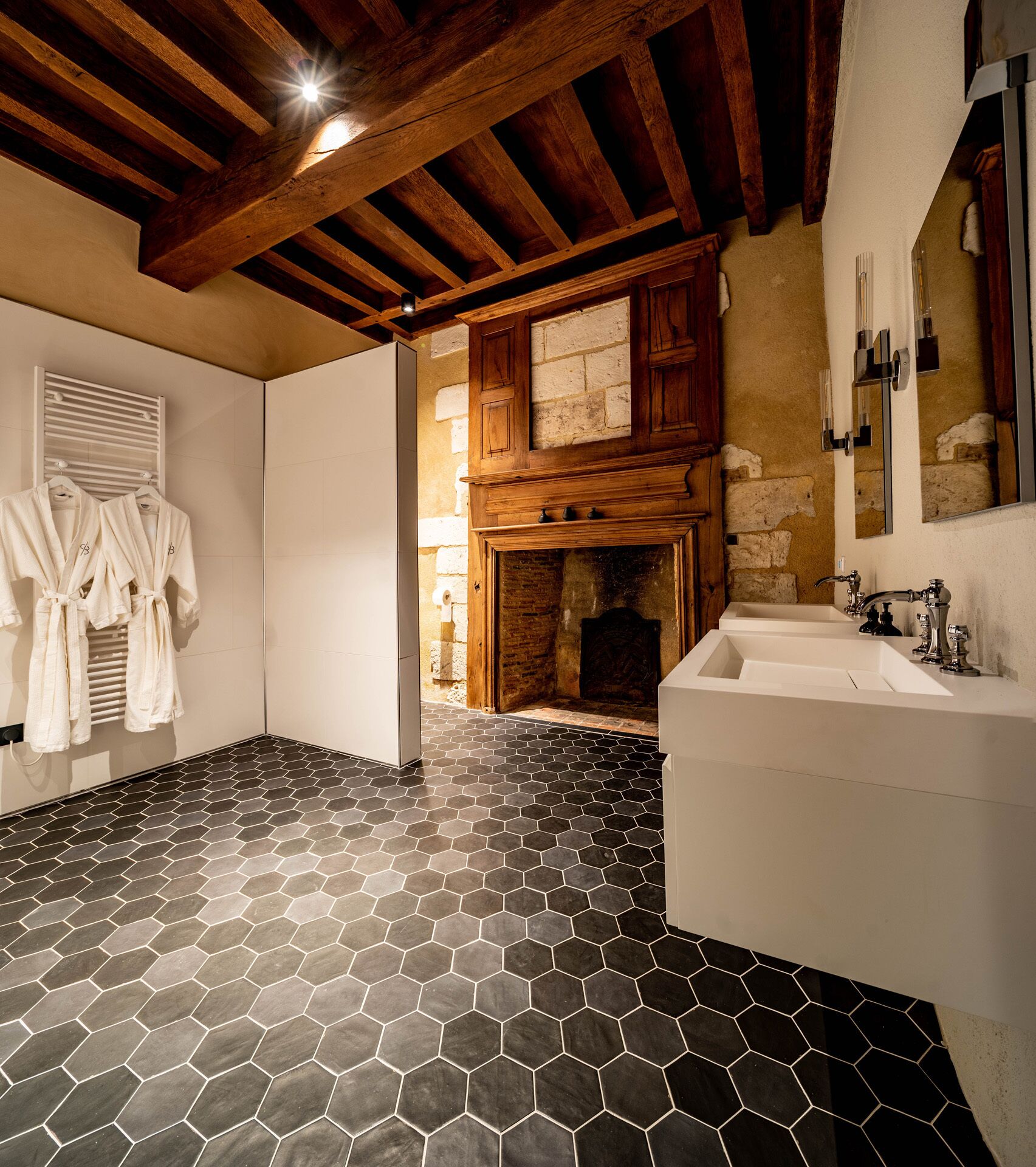Une salle de bain avec un sol carrelé en forme d’hexagone, des poutres en bois, une cheminée et un grand miroir.