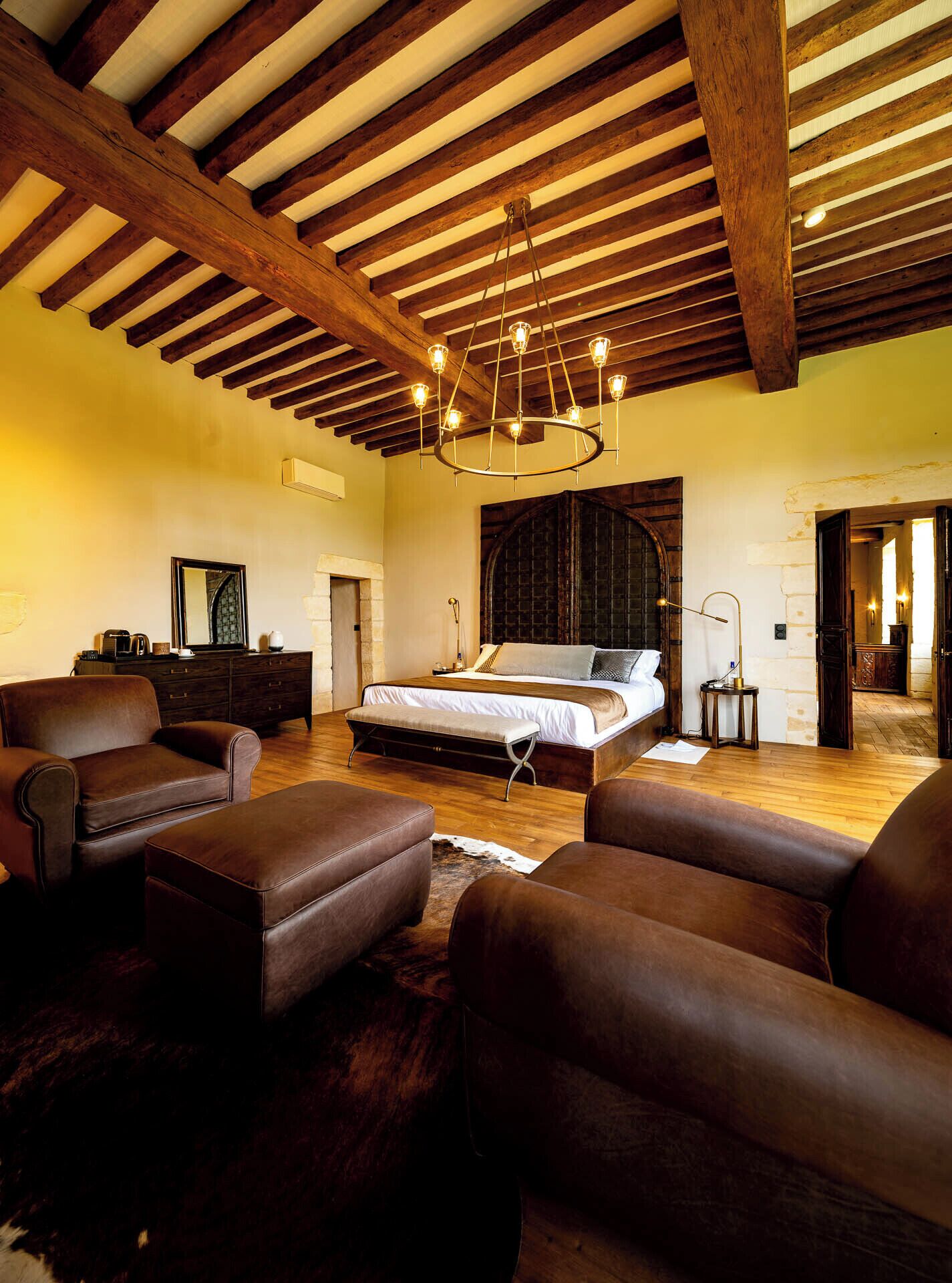 A spacious bedroom with a large bed, wooden beams, a chandelier, leather furniture, and a rustic stone wall.