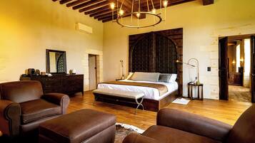 A spacious bedroom with a large bed, wooden beams, a chandelier, leather furniture, and a rustic stone wall.