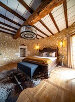 Un dormitorio con una cama grande, vigas de madera, paredes de piedra, una araña y una chimenea de piedra.