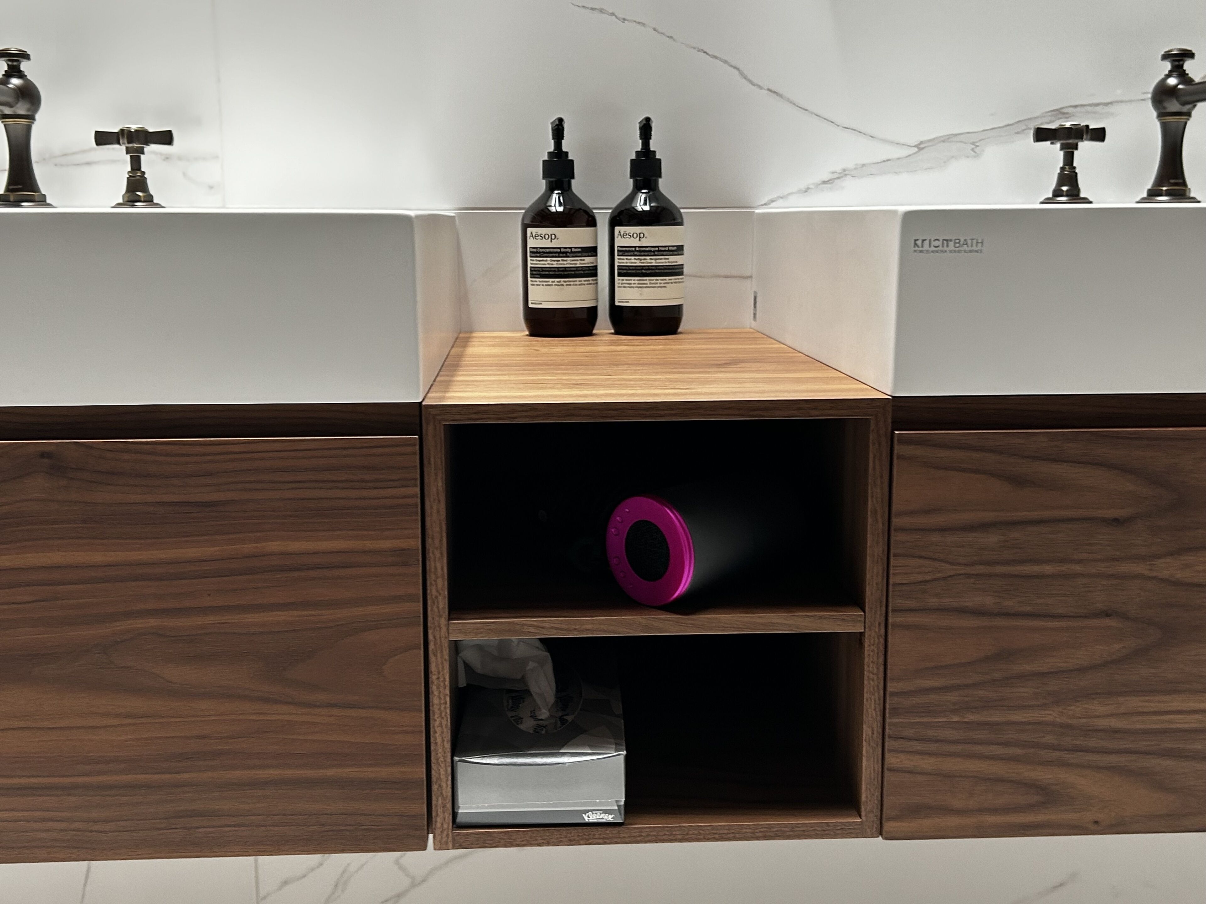 A bathroom vanity with a wooden cabinet, two bottles of Aesop body wash, a pink soap dish, and a small silver box.