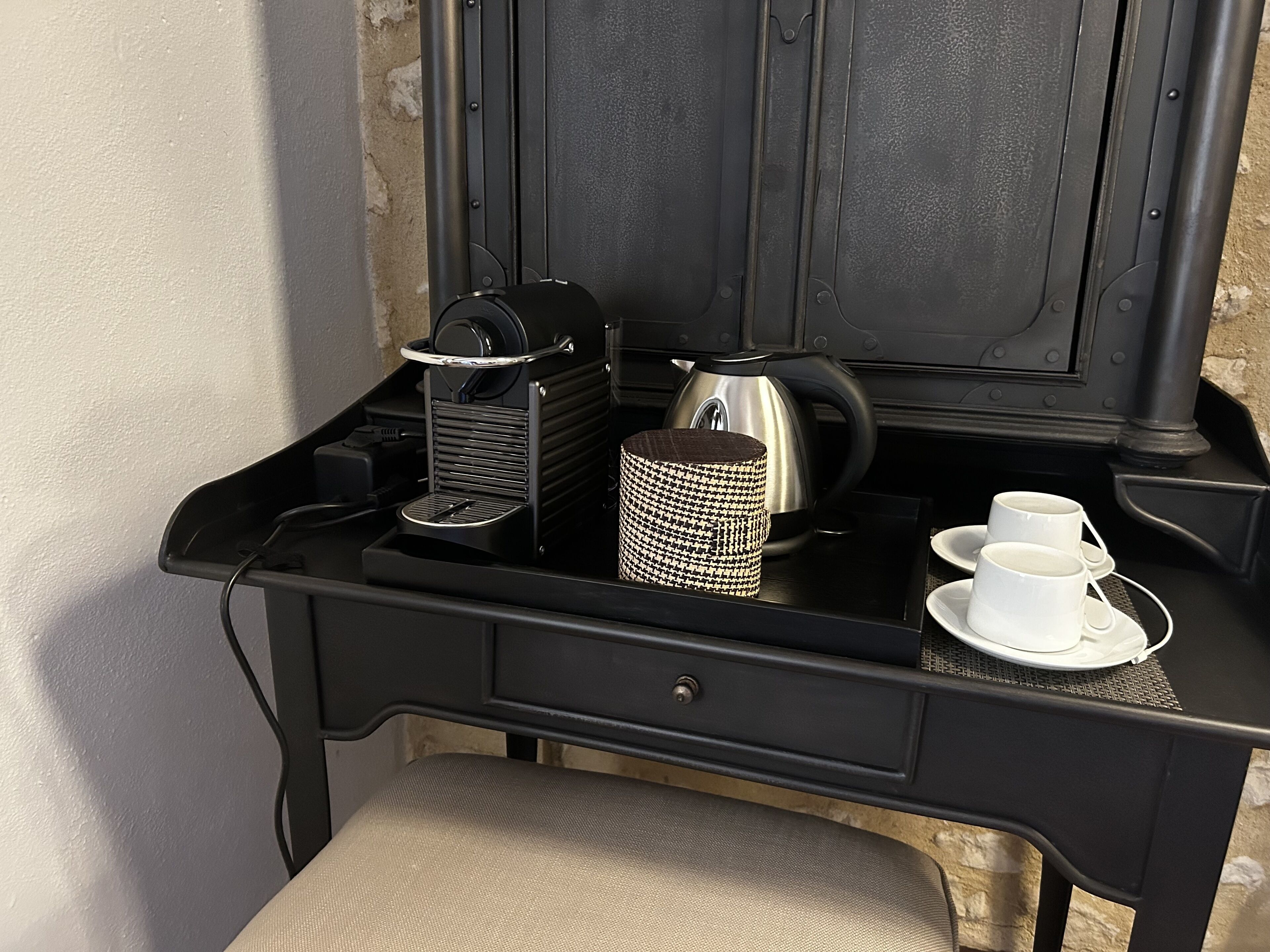 A compact kitchenette with a coffee maker, kettle, and tea set on a black countertop.