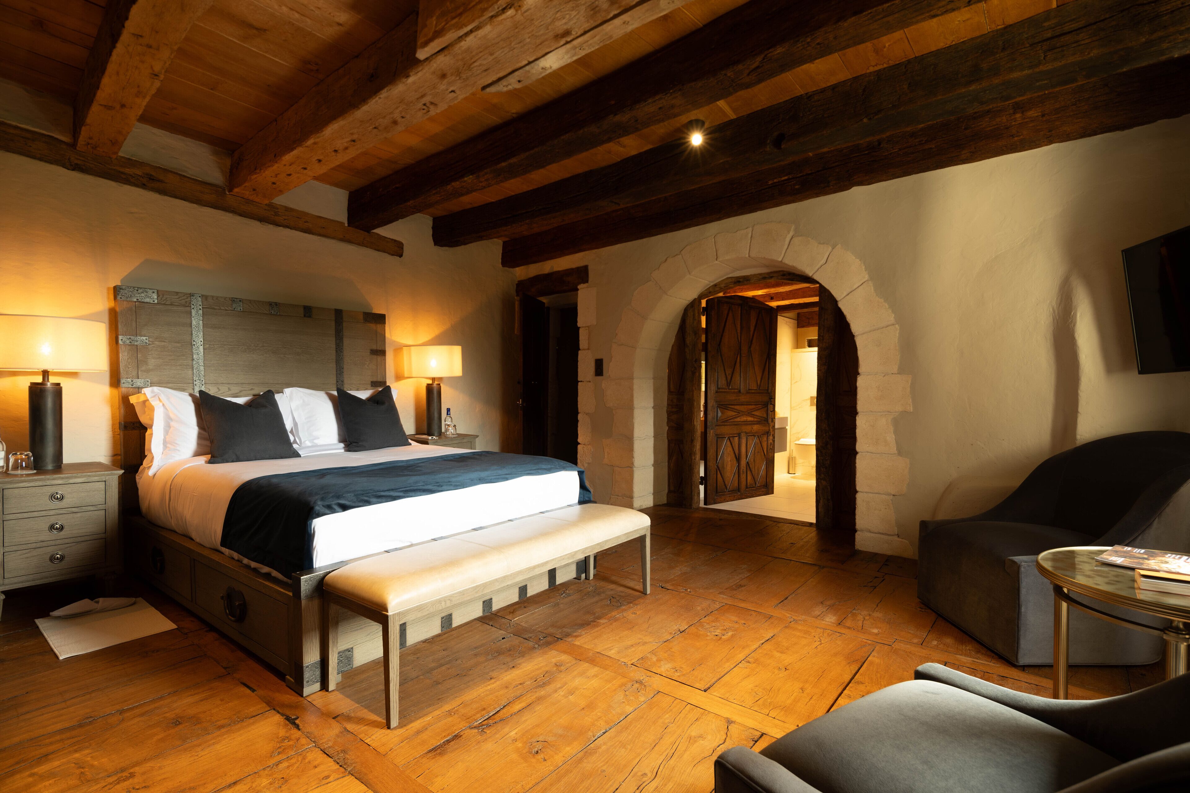 Un dormitorio con una cama grande, suelo de madera, un banco, una silla y un arco de piedra.