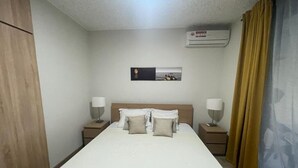 Ett hotellrum med en stor säng, två sängbord, en väggmonterad luftkonditioneringsanläggning och ett fönster med gardiner.