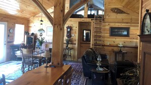 Interior de una cabaña de madera con comedor, sala de estar con sofá y mesa de centro, y una escalera que conduce a un nivel superior.