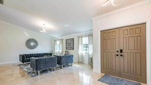 A spacious living room with a large beige carpet, a dark-colored sofa set, a decorative wall piece, and a wooden door.