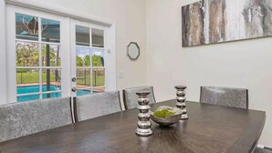 A dining area with a dark wooden table, six chairs, and two decorative candle holders with green moss. A large abstract painting is on the wall, and a window reveals a pool and greenery outside.