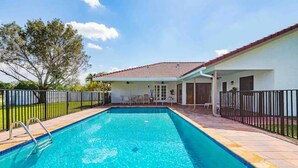 A rectangular swimming pool with a tiled deck, surrounded by a fence, and a covered patio area with chairs and a table.