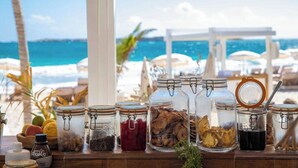 Ein Tisch mit verschiedenen Speisen und Gewürzen, darunter Gläser mit Kräutern und eine Saucenflasche, vor einer Aussicht auf Strand und Meer.