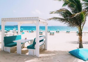 Een zithoek aan het strand, met een witte pergola, groene kussens en een tafel.