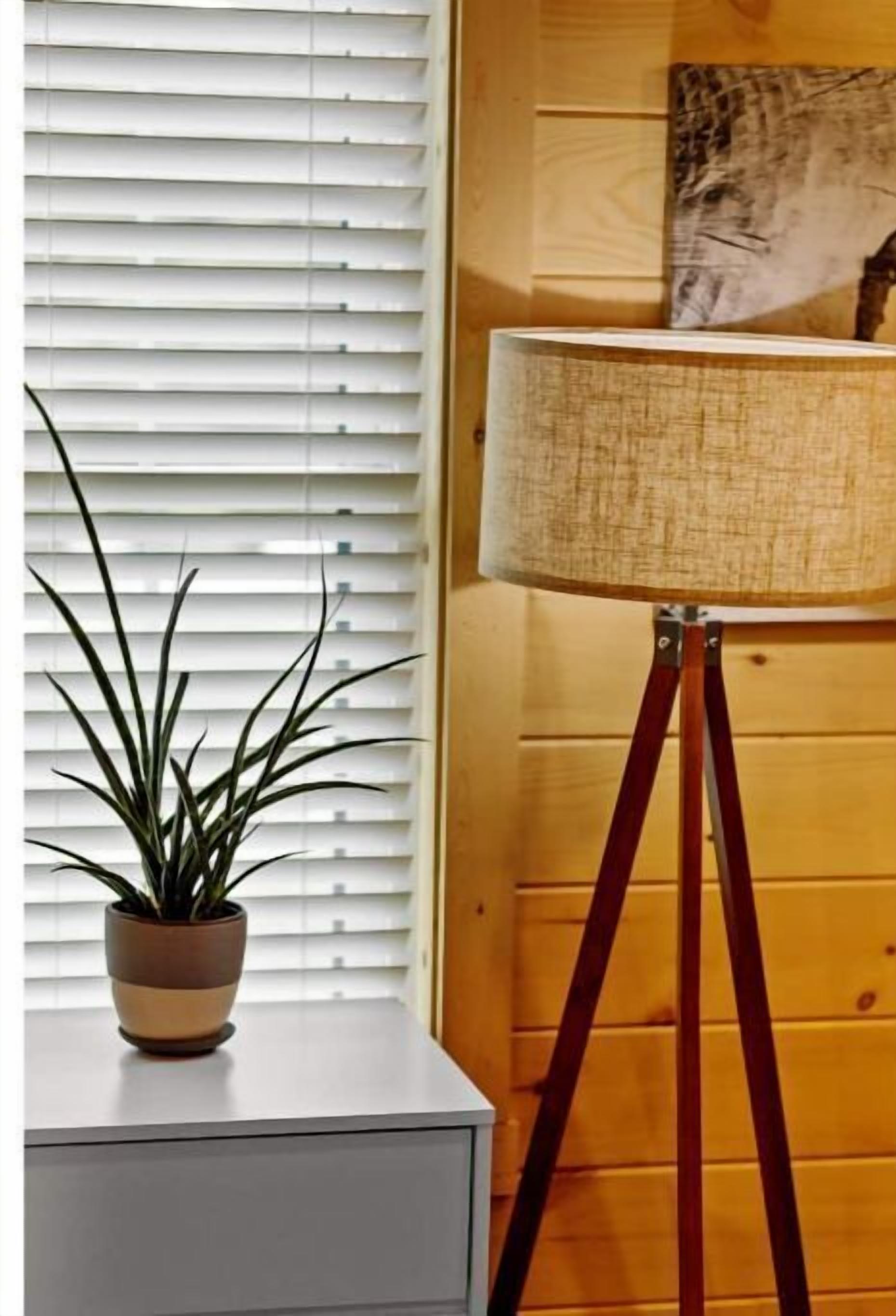 A room with a potted plant on a windowsill, a floor lamp with a textured lampshade, and a wooden wall with a painting.