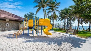 A playground with a yellow spiral slide and climbing structures, set against a backdrop of palm trees and a beach. - Condo 300 steps from beach with heated pool and private free parking. (Pompano Beach)