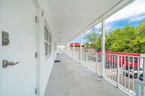 Ein überdachter Balkon mit weißer Geländer, eine Tür mit Griff und Blick auf geparkte Autos sowie ein Gebäude mit rotem Dach.