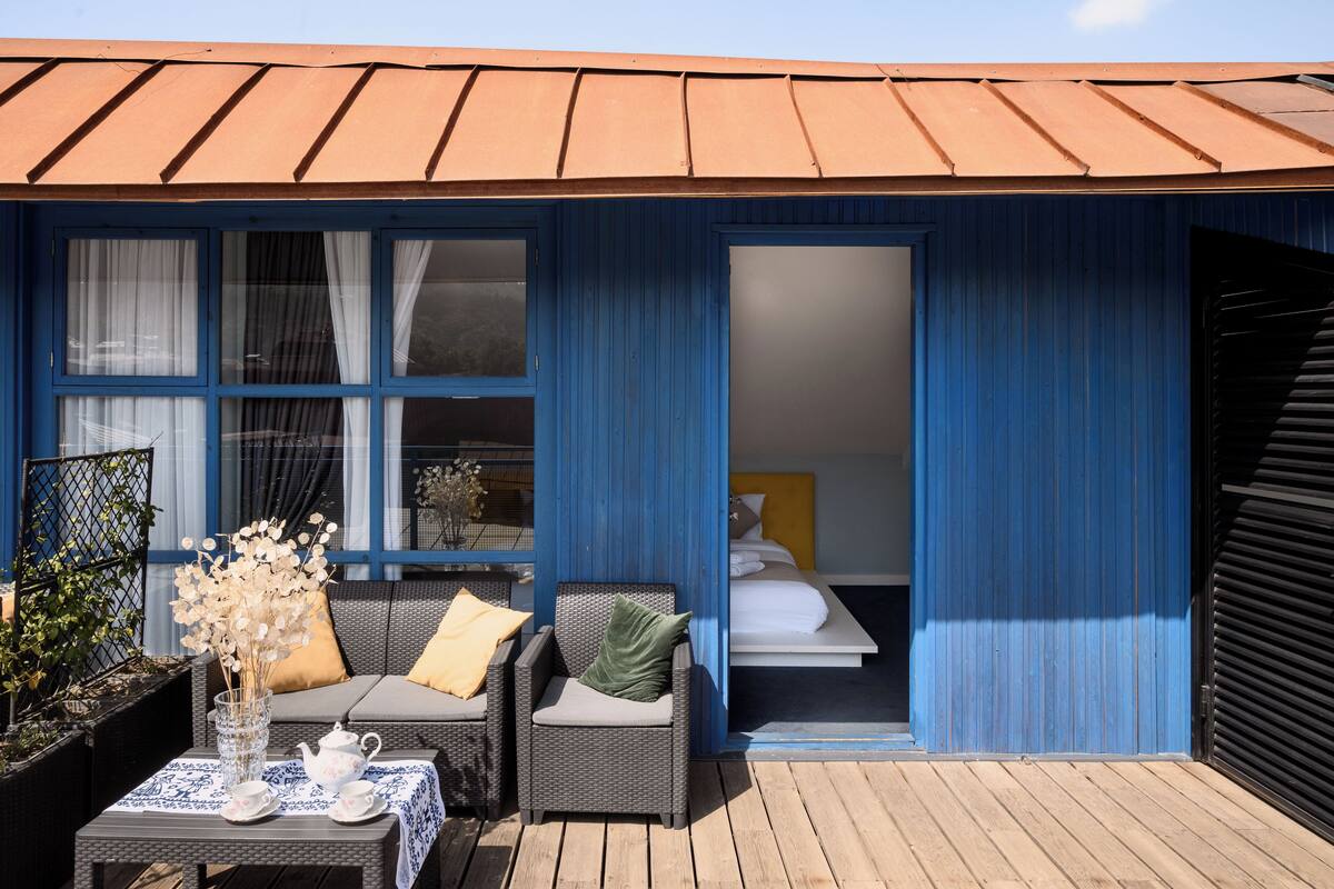 A blue exterior with a wooden deck, a wicker sofa with cushions, a small table with a vase of flowers, and a glimpse of a bed inside.
