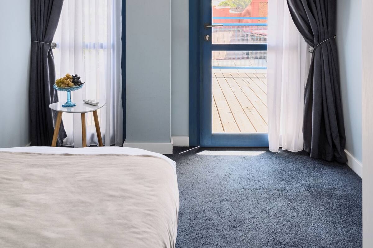 A hotel room with a bed, a small table with a bowl of fruit, a TV mounted on the wall, and a balcony with a view of buildings.