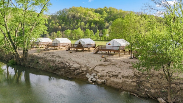 Un camping con varias tiendas de campaña sobre pilares en una zona arenosa cerca de un río, rodeado de árboles.