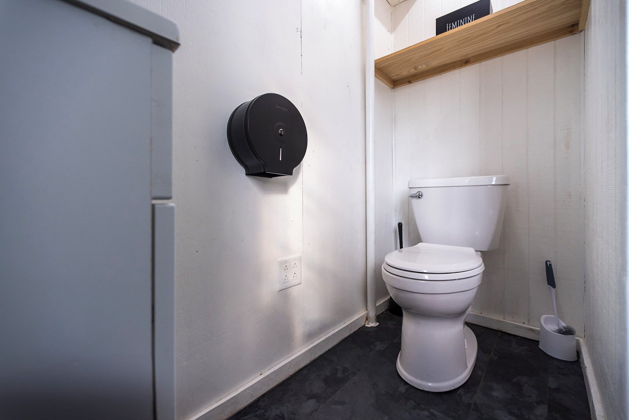 A bathroom with a toilet, a black toilet paper holder, a broom, and a shelf with a sign.