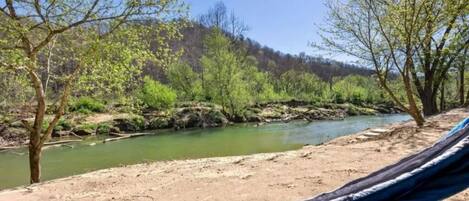 A hammock on a sandy beach with a river and trees in the background.