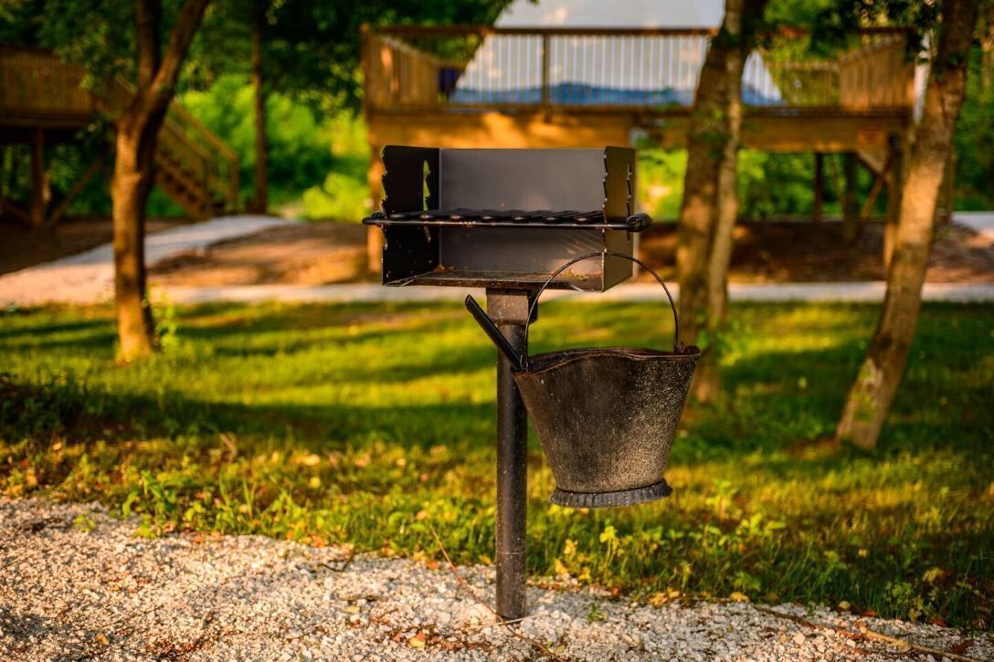 Una parrilla para barbacoa con un cubo metálico unido a un lado, sobre una superficie de grava con césped y árboles al fondo.
