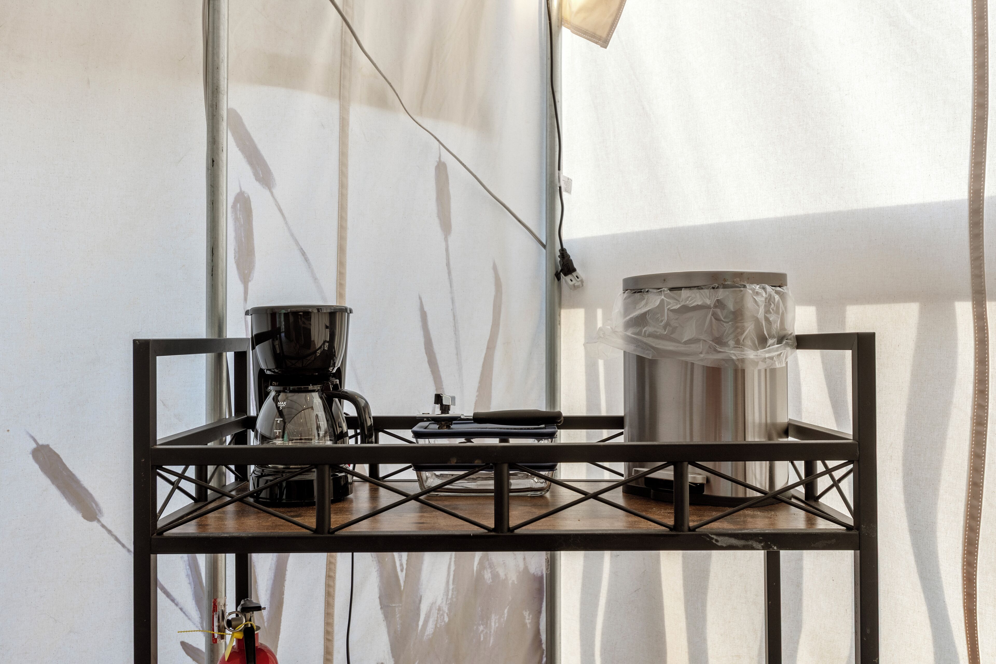 Una mesa de estructura metálica con una cafetera y un recipiente, posiblemente para agua o té, debajo de una estructura tipo tienda.