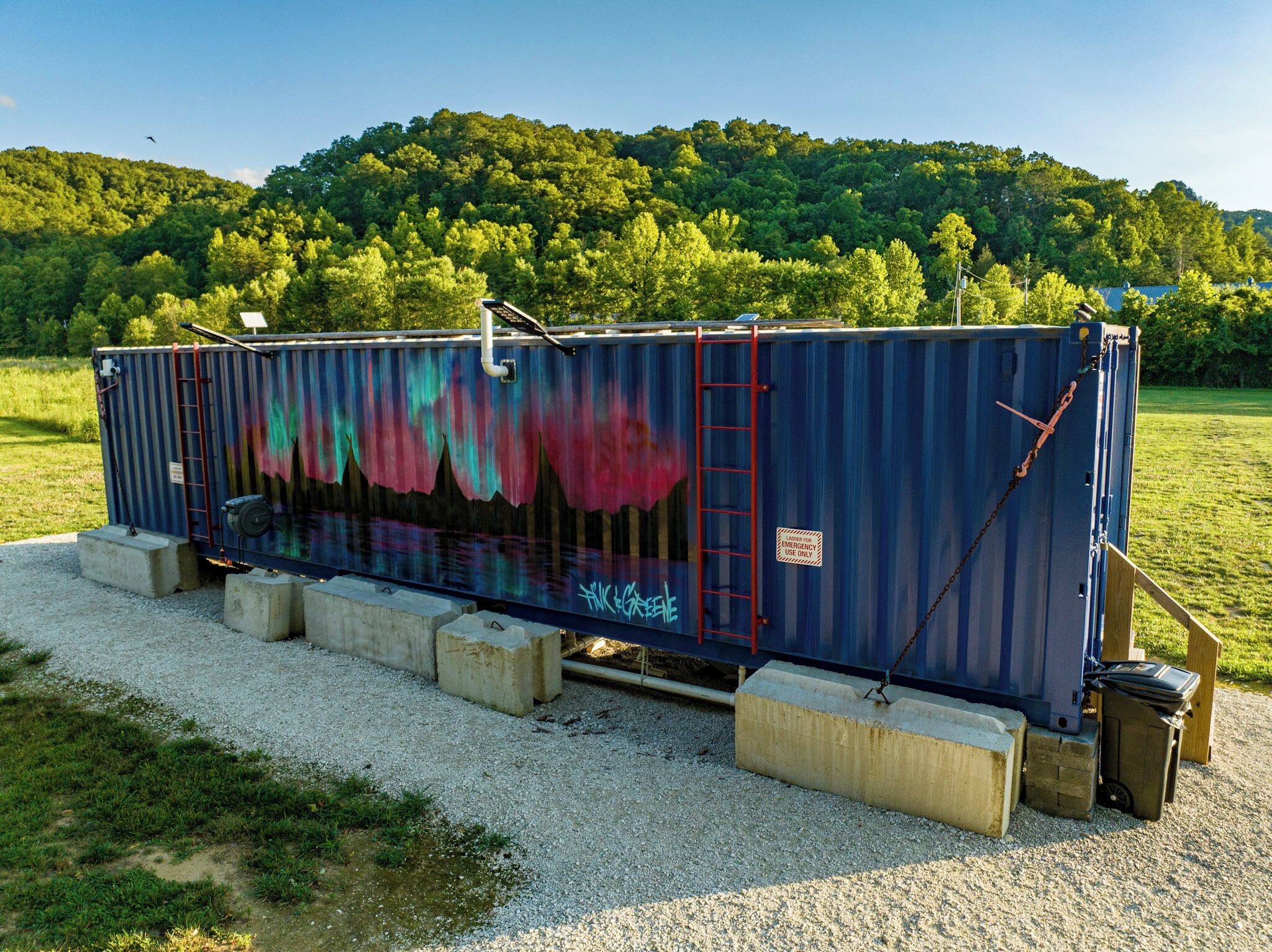 A large shipping container with colorful graffiti, secured with chains and a ladder, placed on concrete blocks in a grassy area with a forested hill in the background.
