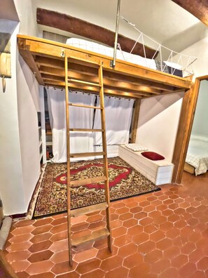 Ein Zimmer mit einem Holzbett, einer Himmelsleiter, einem gemusterten Teppich und einem sechseckig gefliesten Boden.