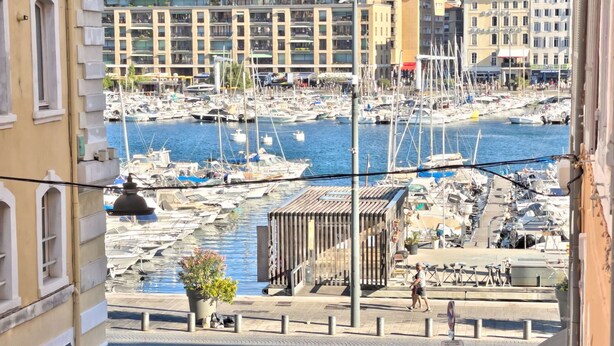 Un puerto deportivo con barcos amarrados, un paseo peatonal y edificios al fondo.