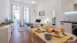 Een moderne, goed verlichte kamer met een ontbijtgedeekte eethoek, een tafel met borden eten en drinken, een bed met een blauwe hoofdbord en een keuken met een spoelbak en kastjes.
