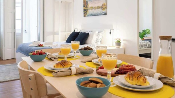 Een ontbijt met croissants, fruit en sinaasappelsap op een houten tafel in een lichte kamer, met een bed en een schilderij op de achtergrond.