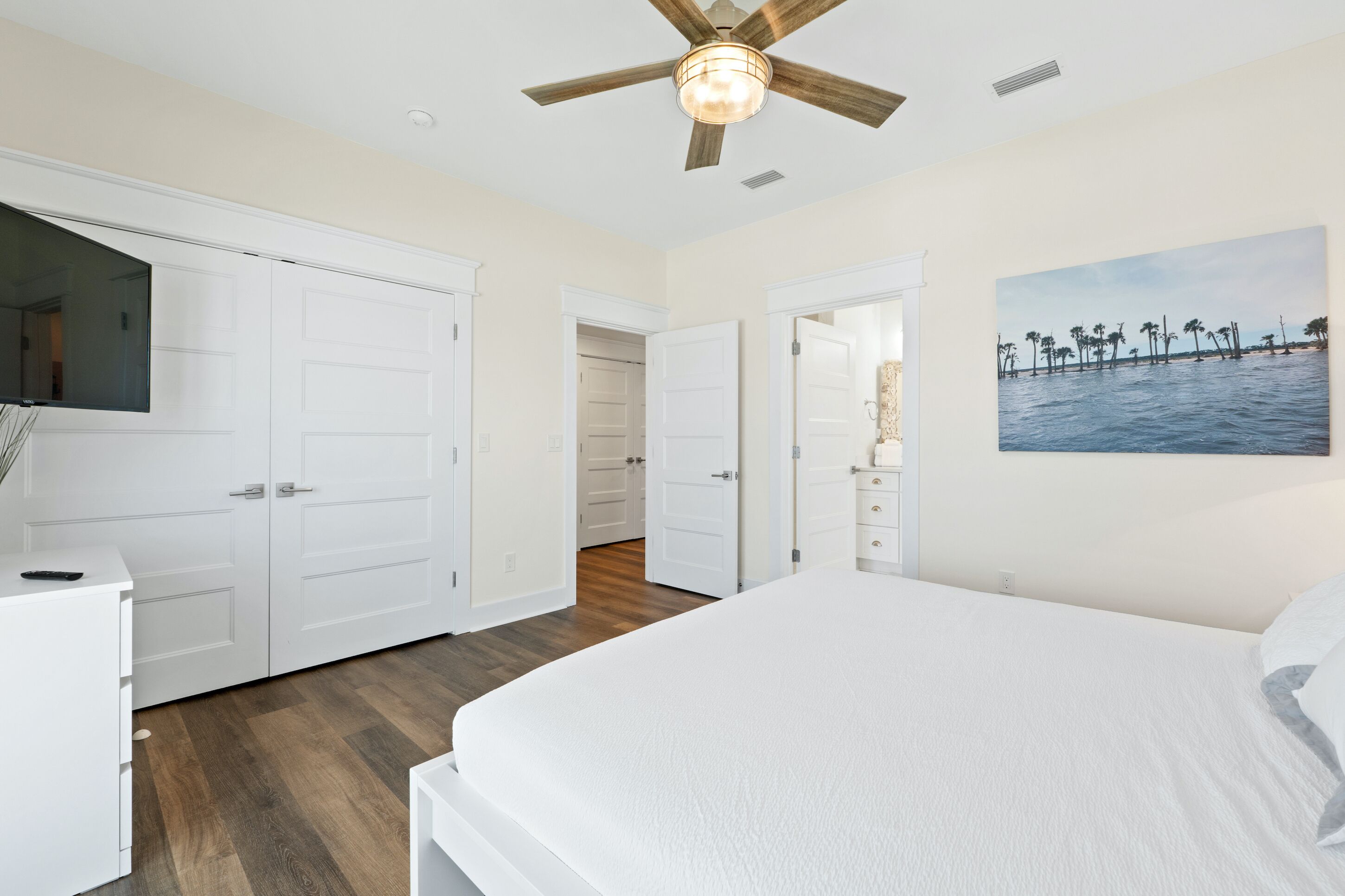 A bedroom with a large bed, a ceiling fan, white wardrobes, and a painting of palm trees on the wall.