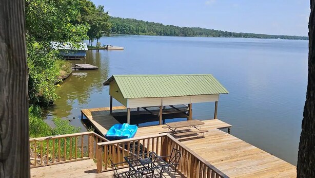 A wooden deck with a covered seating area overlooking a calm lake, surrounded by lush greenery. - Lakeside Chalet on Beautiful Gantt Lake! (Dozier)