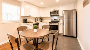 A modern kitchen with white cabinets, stainless steel appliances, a round wooden table, and chairs. - (12) Book Now & Save 10%! Modern July Apartment (Houston)