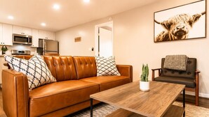 A living room with a brown leather sofa, a wooden coffee table, a potted plant, a framed picture on the wall, and a kitchen in the background. - (12) Book Now & Save 10%! Modern July Apartment (Houston)