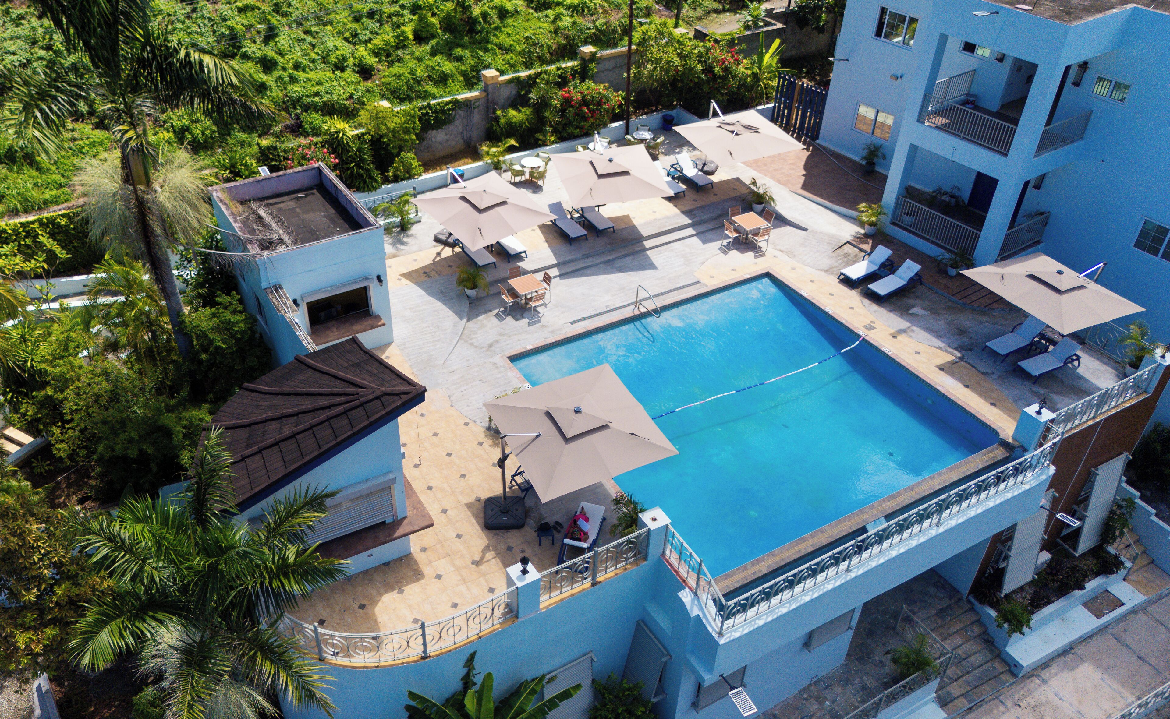 Vista aérea de un hotel con piscina rodeada de sillas de descanso y sombrillas, un espacio de terraza cubierto y abundante vegetación.