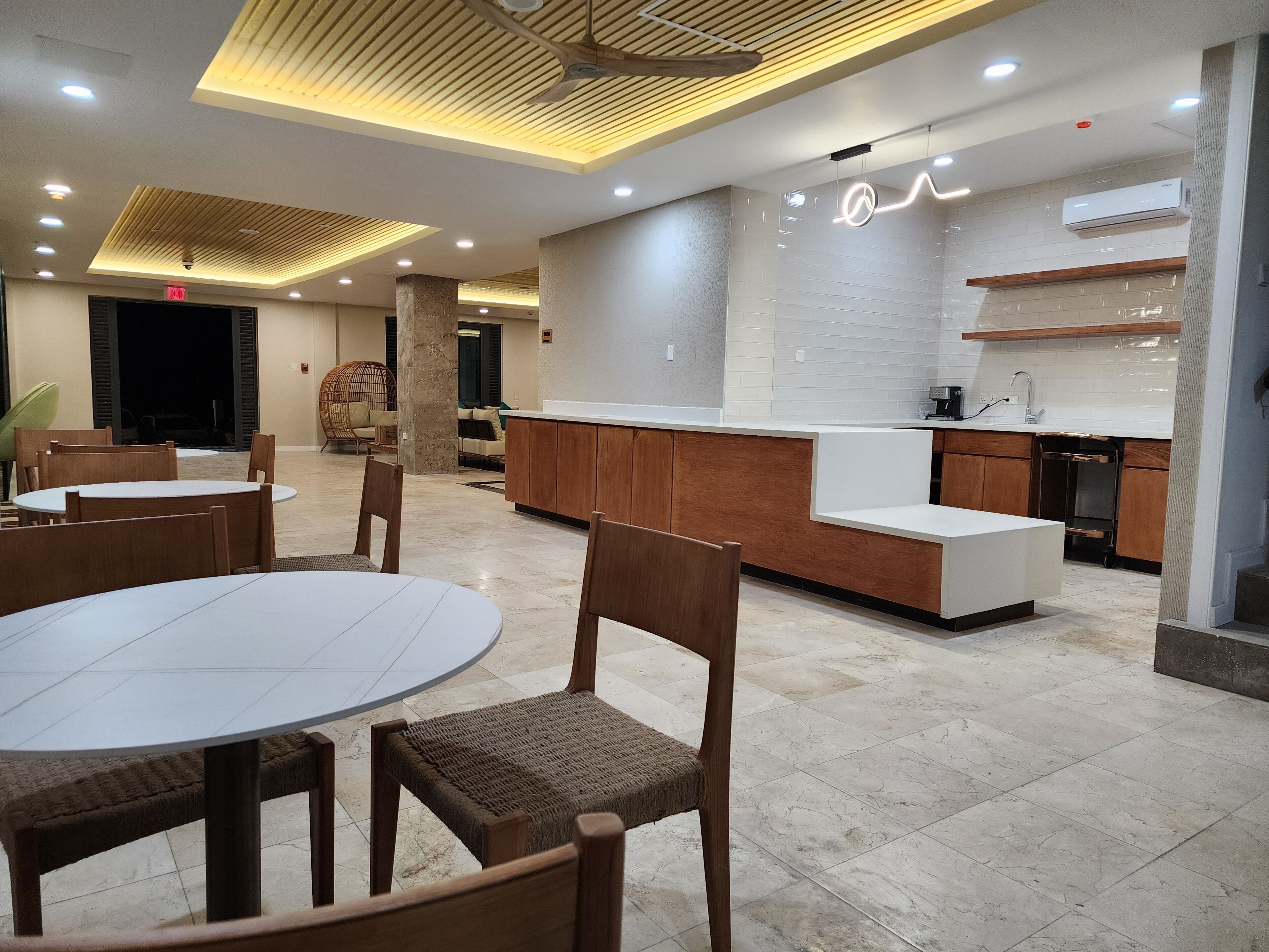 Een moderne hotel lobby met ronde tafels en houten stoelen, een receptiebalie en een keukenhoek.