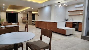 Een moderne hotel lobby met ronde tafels en houten stoelen, een receptiebalie en een keukenhoek.