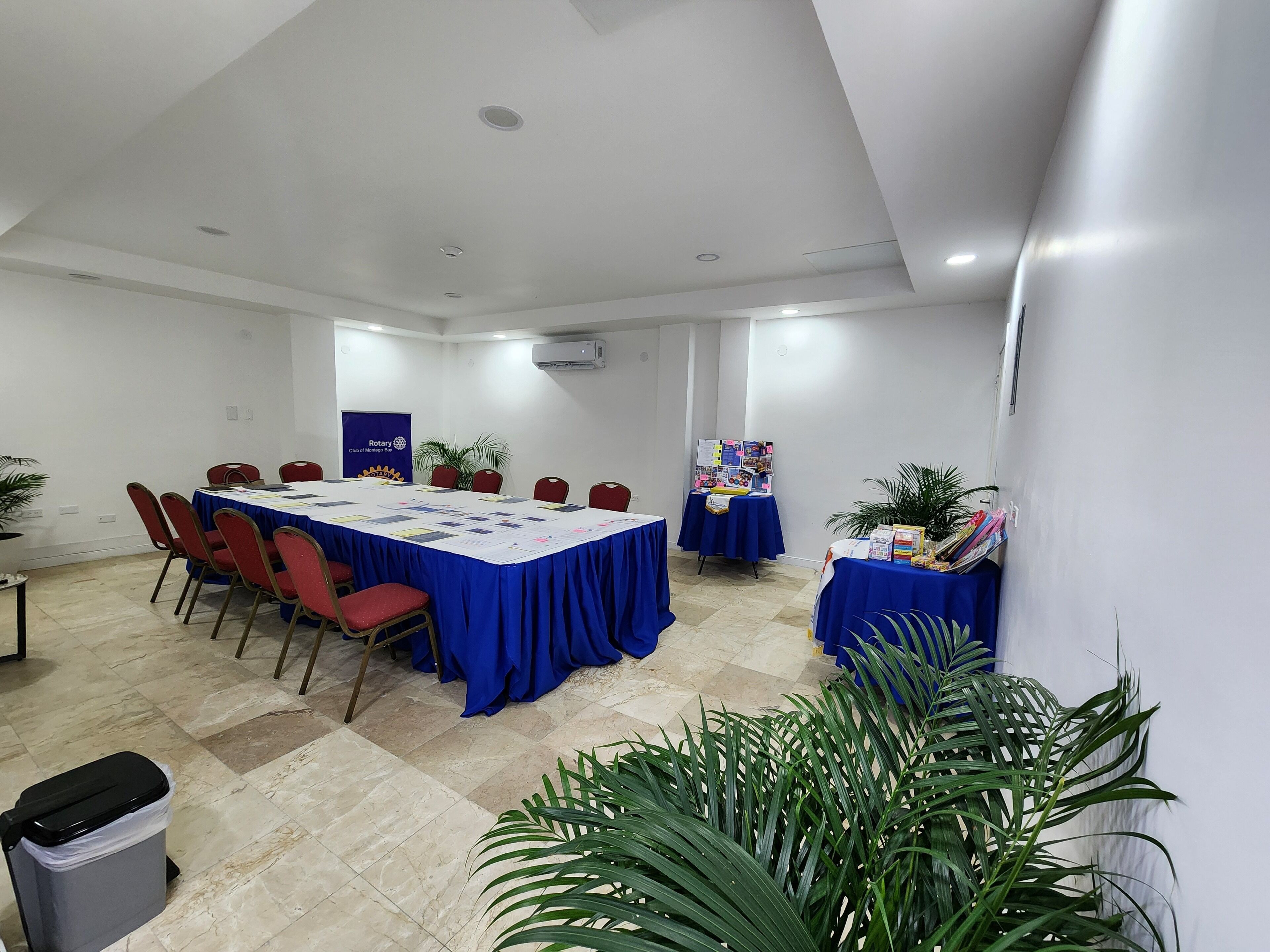 Un salón de conferencias con una larga mesa, sillas rojas y un mantel azul. Hay plantas en macetas y un tablón de anuncios con papeles.