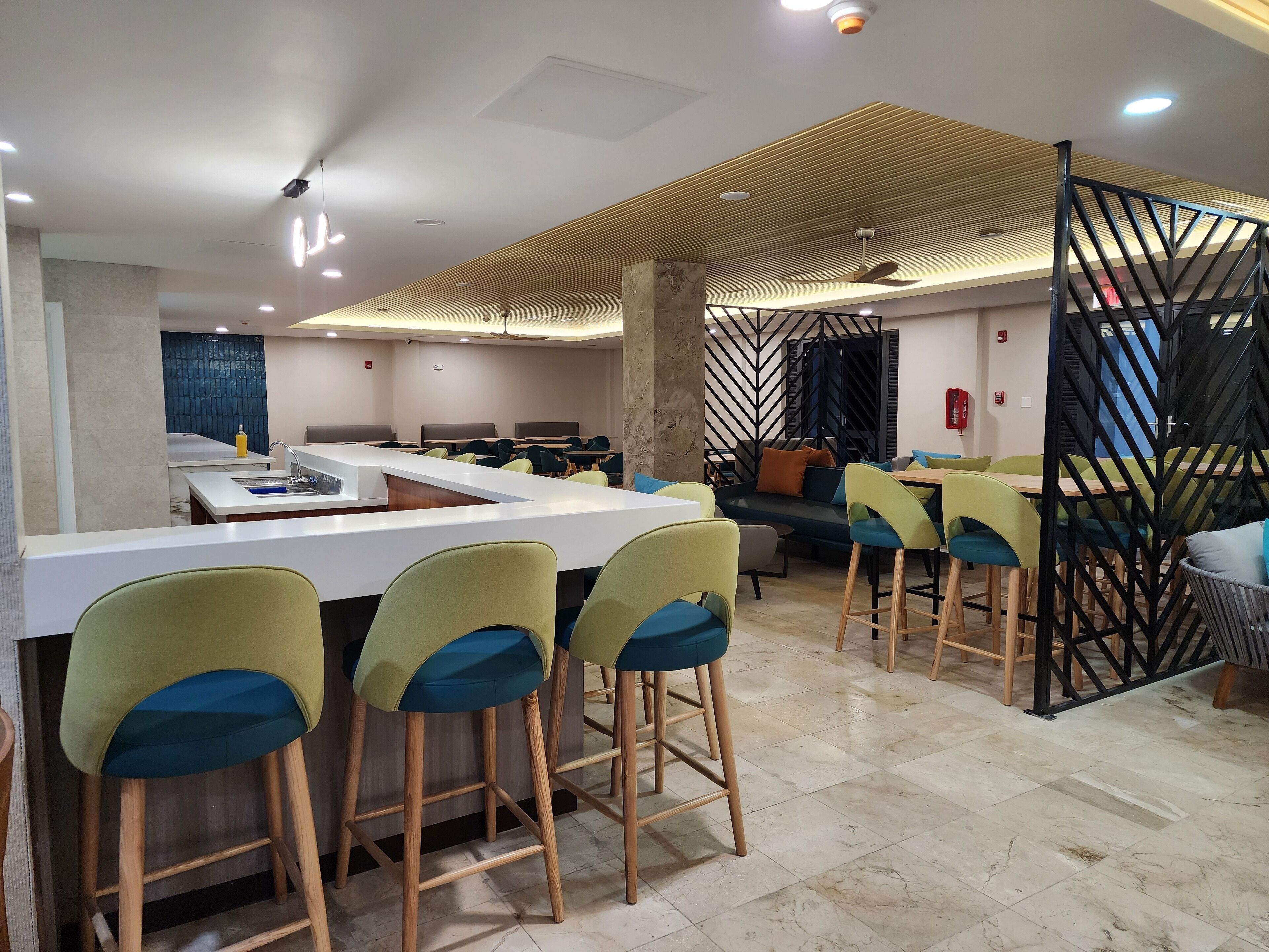 A modern restaurant with a central bar area, high ceiling with recessed lighting, and a herringbone-patterned ceiling.