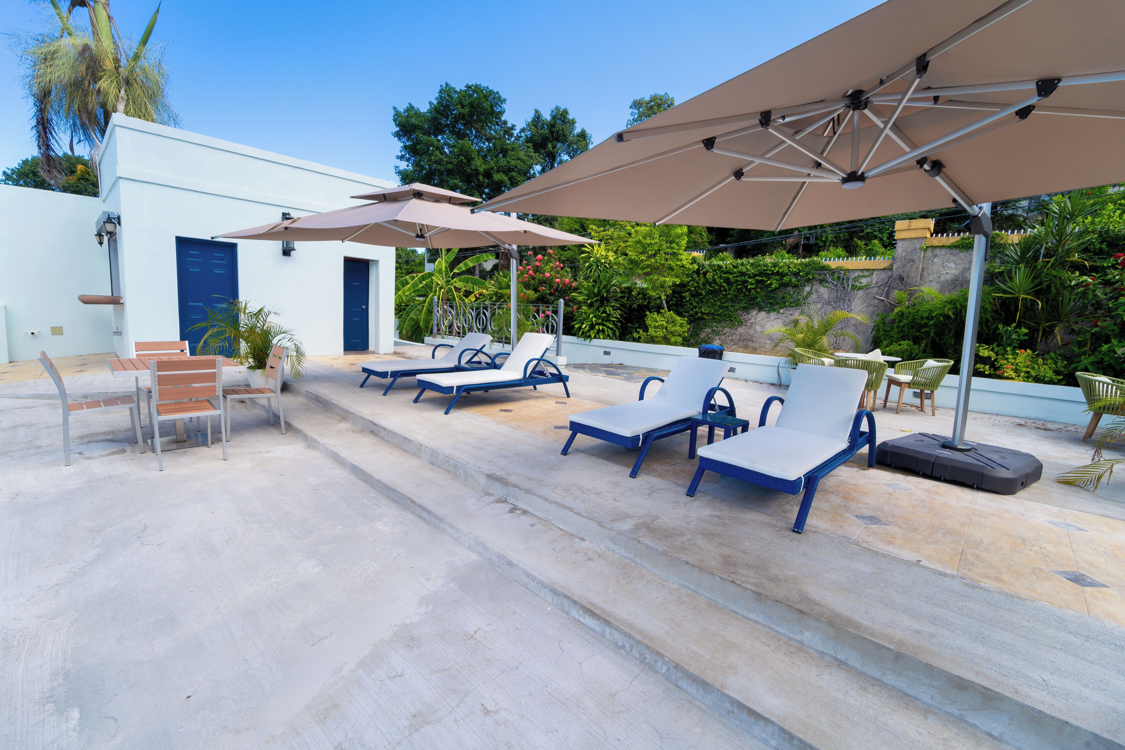 An outdoor seating area with lounge chairs and umbrellas, a white building, and lush greenery in the background.
