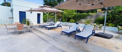An outdoor seating area with lounge chairs and umbrellas, a white building, and lush greenery in the background.