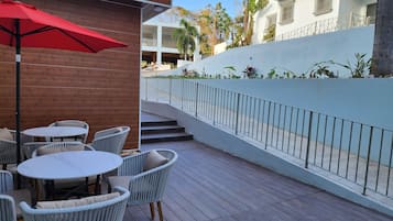 Une terrasse extérieure avec des chaises en osier et un parasol rouge, une plateforme en bois et un mur blanc agrémenté d’une rambarde en métal.