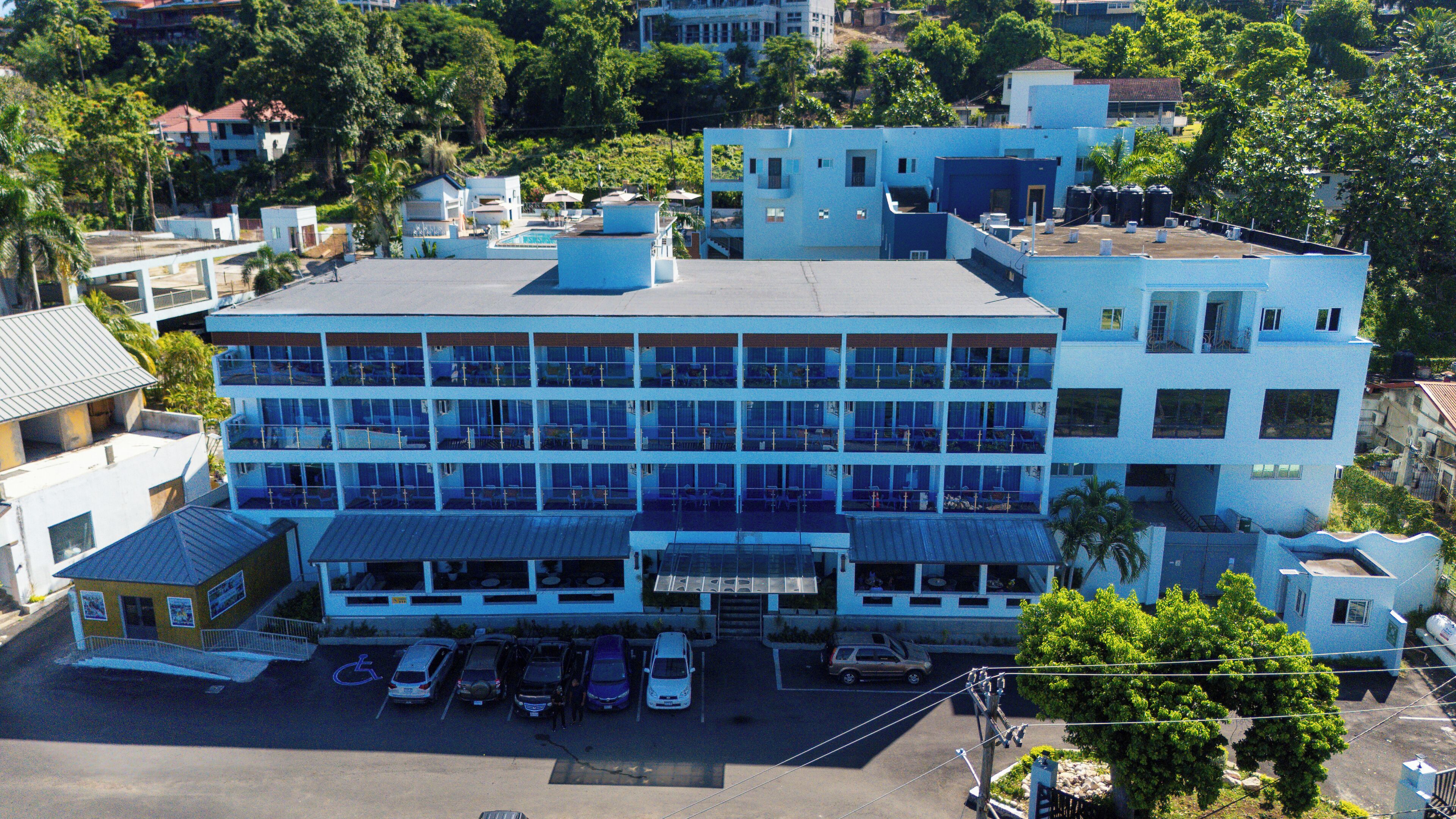 Ein mehrstöckiges Hotel mit blauer Fassade, ein Parkplatz mit mehreren Autos und eine grüne Umgebung.