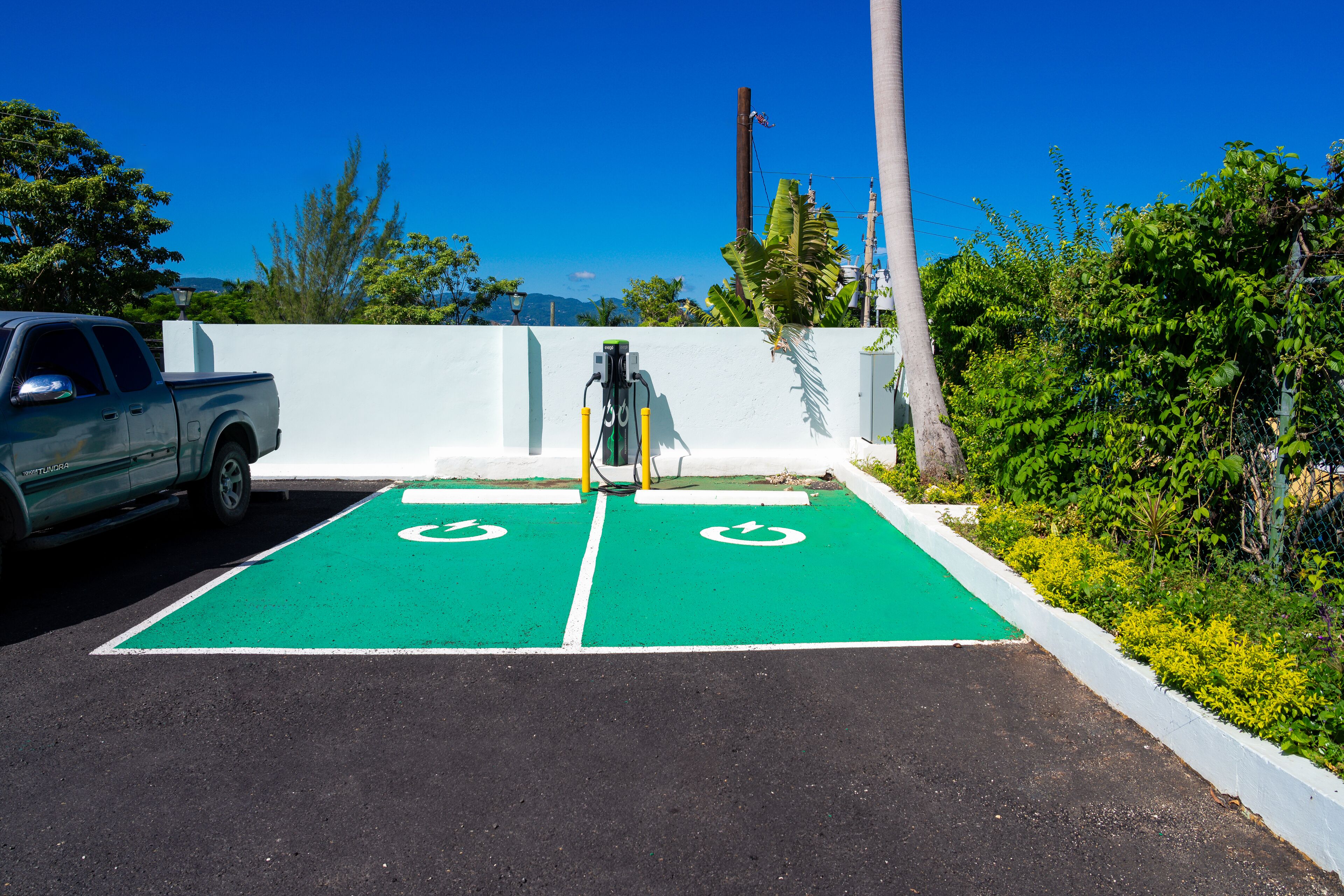 A parking area with electric vehicle charging stations, a white wall, and greenery.