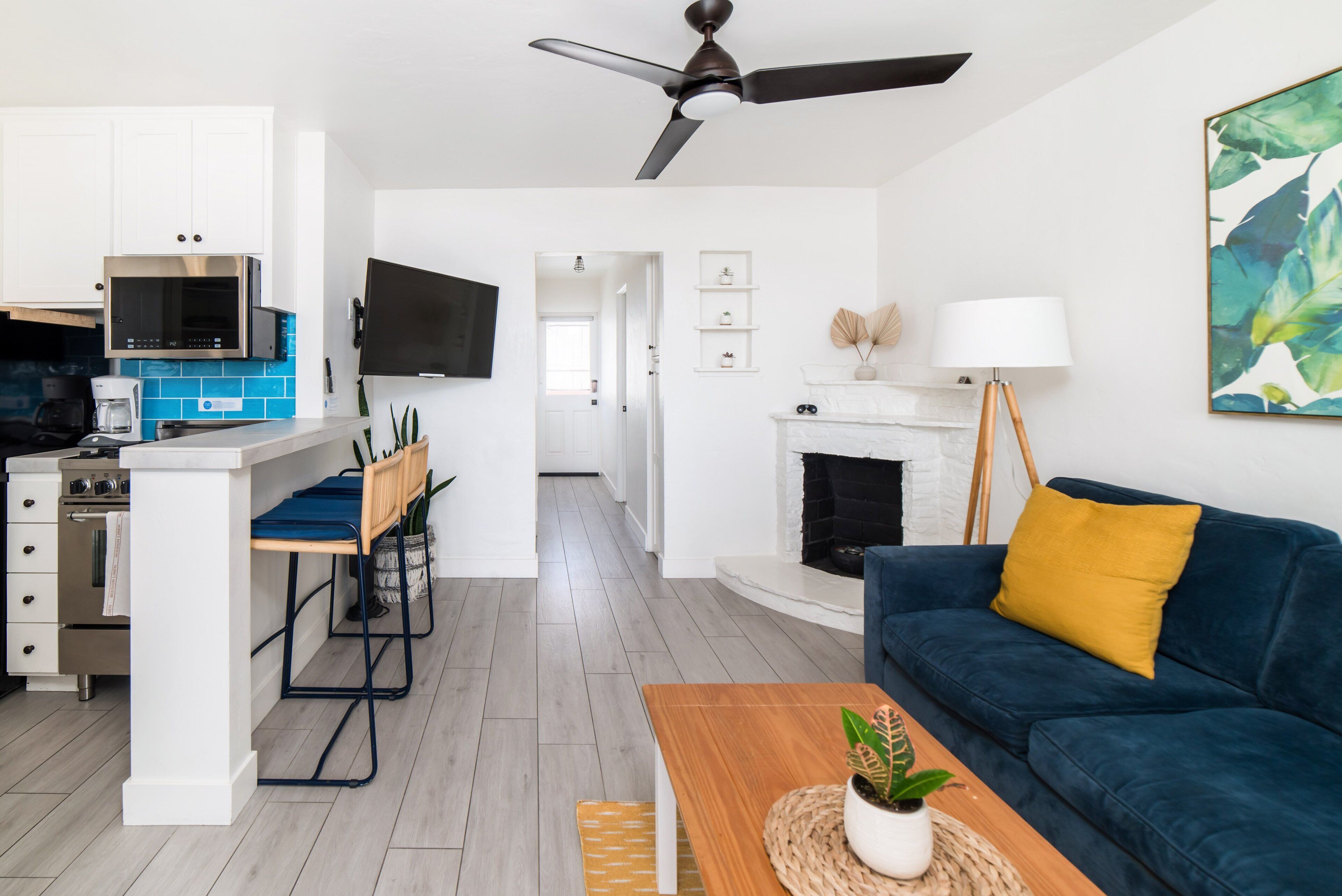 Una cocina moderna con armarios blancos, un microondas integrado y una cafetera. Hay una zona de comedor con una mesa y sillas de madera. Un sofá azul y un cojín amarillo están en la sala de estar. Se ve una chimenea al fondo.