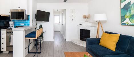 Una cocina moderna con armarios blancos, un microondas integrado y una cafetera. Hay una zona de comedor con una mesa y sillas de madera. Un sofá azul y un cojín amarillo están en la sala de estar. Se ve una chimenea al fondo.