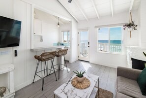 A bright, open living area with a kitchen, a dining area with bar stools, a large TV, and a view of the ocean through a sliding glass door. - The Pacific Villas (Oceanside)