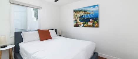 A bedroom with a bed, a ceiling fan, a painting, a window, and a bedside table.