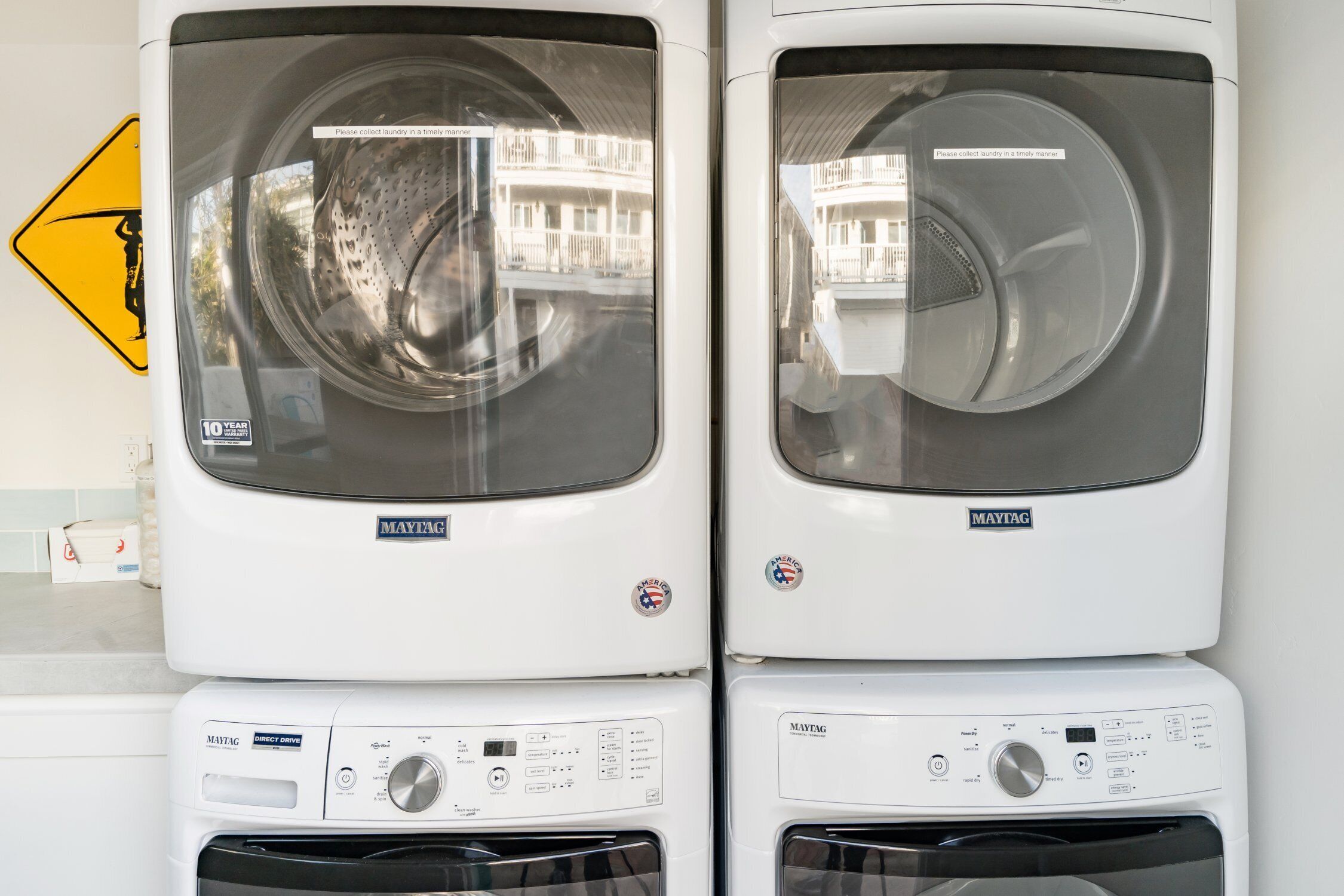Two stacked Maytag washing machines with control panels and a caution sign on the wall.