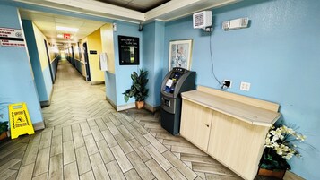 A hotel reception area with a blue wall, a reception desk, an ATM, and a hallway leading to rooms.